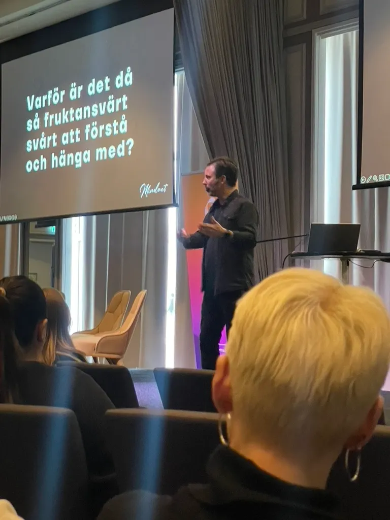 Mathias på scen i en balsal framför sliden ”Varför är det då så fruktansvärt svårt att förstå och hänga med?”