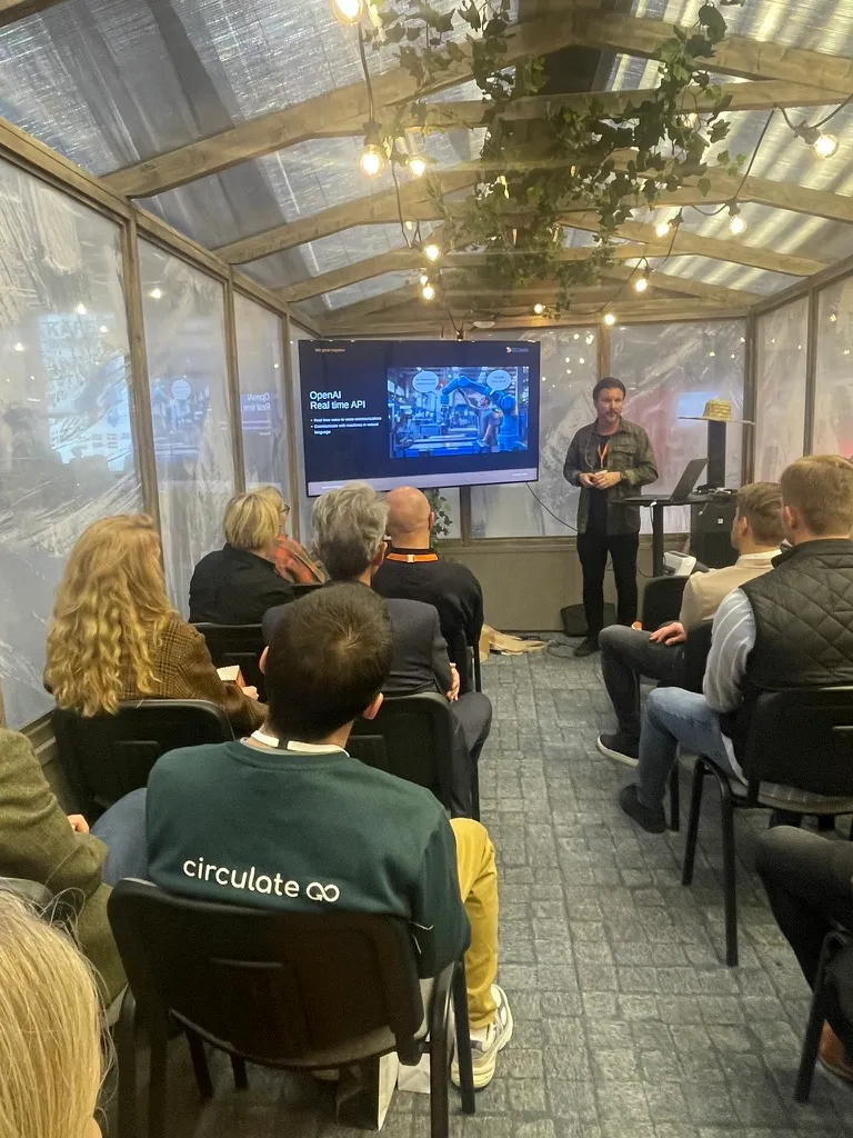 Mathias föreläser i ett växthus med slingerväxter i taket, framför en skärm om OpenAI:s realtids-API